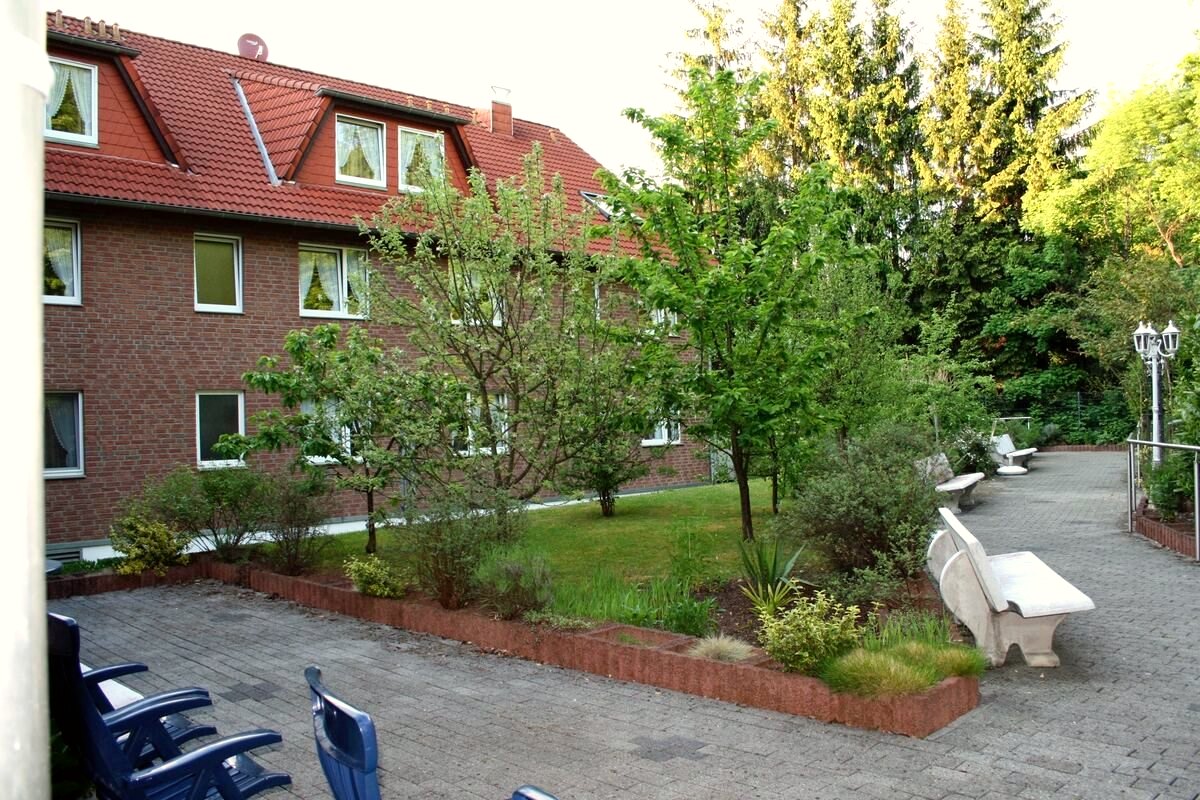 Grünanlage mit Bäumen, Rasenflächen, Wegen und Sitzbänken hinter dem Haus